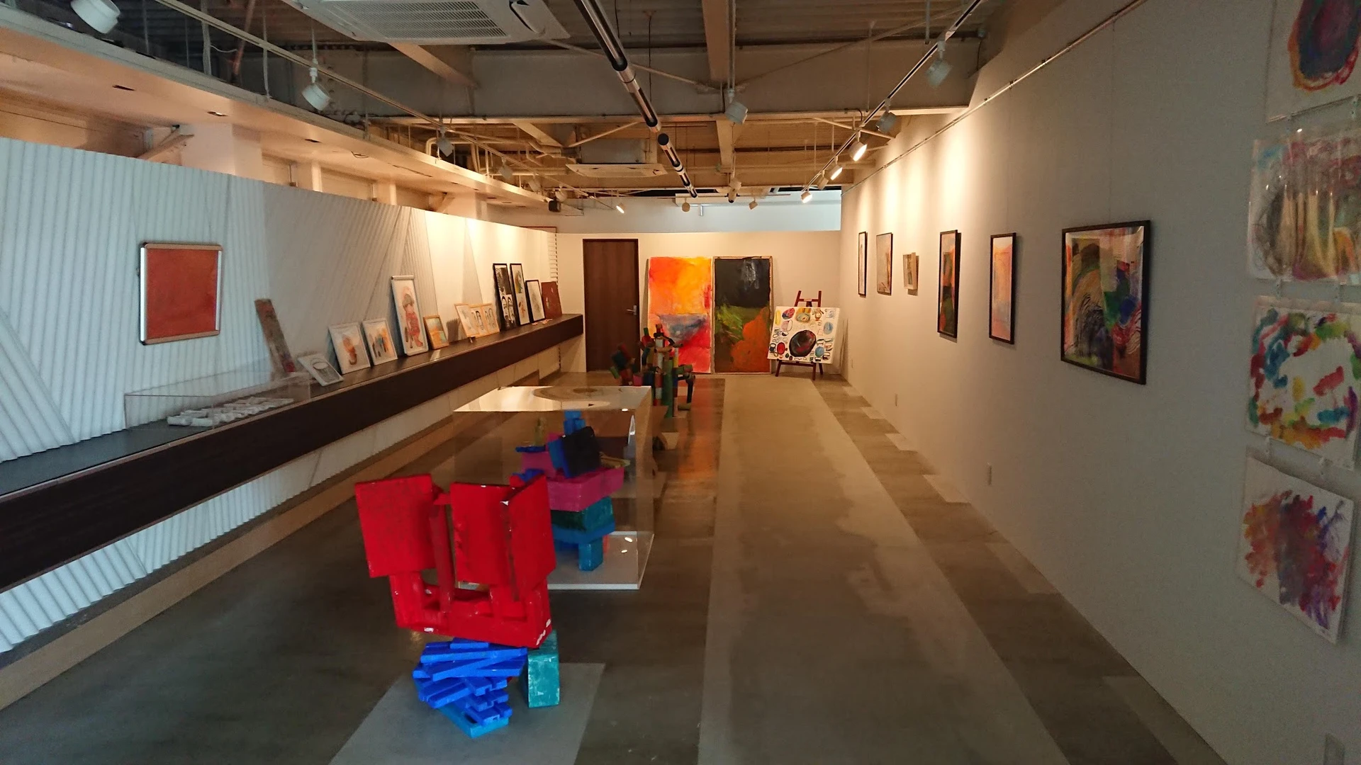 Kunstgalerij-achtig interieur met rode en blauwe sculpturen, schilderijen aan de wanden en geïndustrialiseerd plafond
