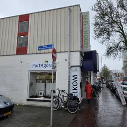 Witte gevel van kringloopwinkel met rode raamdetails, naam PortAgora zichtbaar, fietsen voor de deur