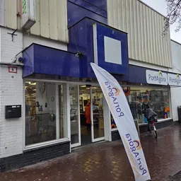 Voorgevel van kringloopwinkel met blauwe luifel, winkelnaam op raam, en personen op nat plein.