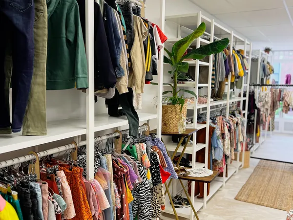Winkelbinnenkant met witte stellingen vol kleding, schoenen en accessoires, een plant in mand, houten vloer