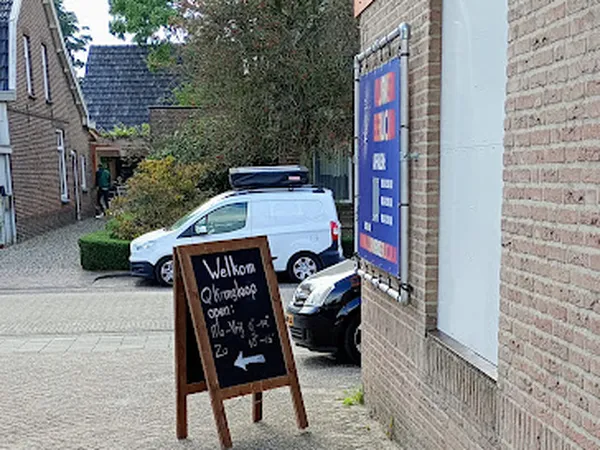 Voorkant kringloopwinkel met houten welkomsbord, blauwe winkelpoort en geparkeerde auto's