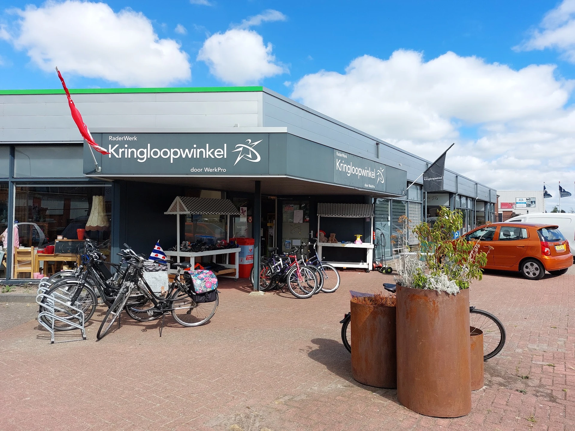 Gevel van kringloopwinkel met naam Kringloopwinkel op donkergroene luifel, met fietsen en spullen buiten, rode auto rechts, b