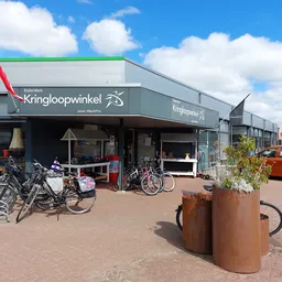 Gevel van kringloopwinkel met naam Kringloopwinkel op donkergroene luifel, met fietsen en spullen buiten, rode auto rechts, b