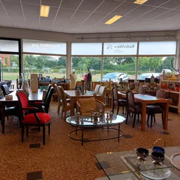Ruim interieur van kringloopwinkel met houten meubels, rode stoelen en grote ramen met uitzicht op groen landschap