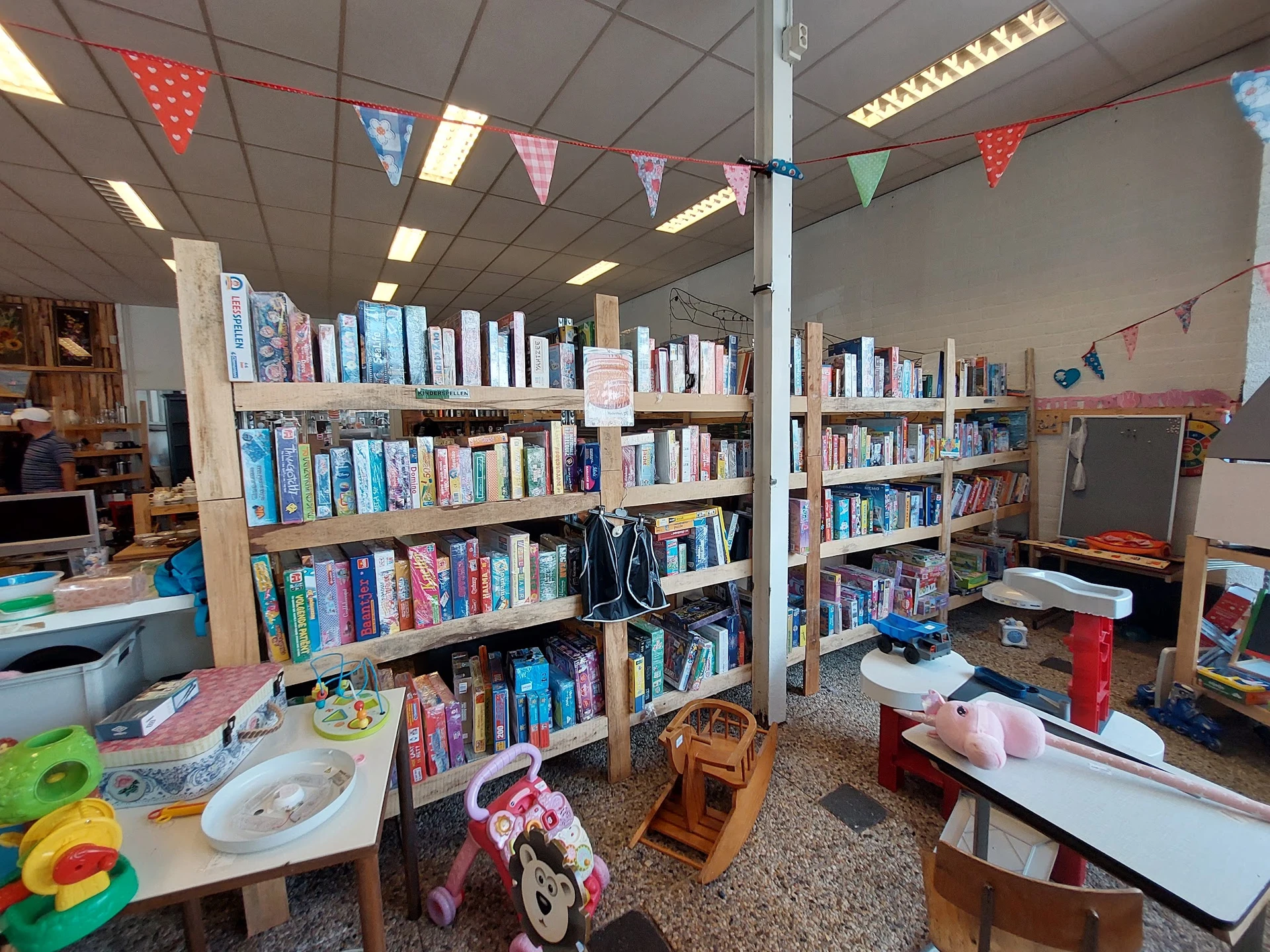 Binnenkant van kringloopwinkel met boekenkasten, speelgoed en kinderafdeling met gekleurde vlaggetjes als versiering
