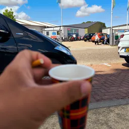 Hand met koffiekopje op parkeerterrein met auto's en bedrijfsgebouwen op achtergrond