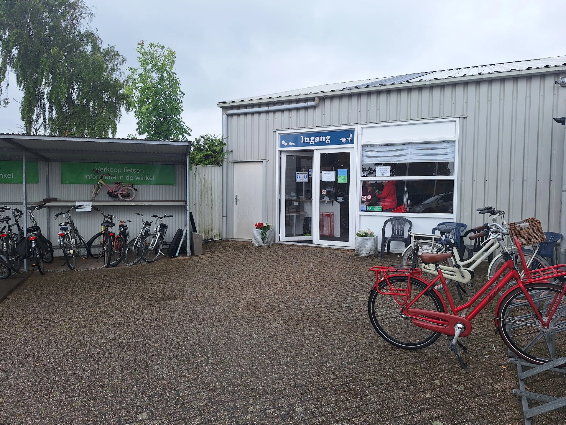 Voorkant van kringloopwinkel met ingang, ramen en uitgestalde fietsen op het terras