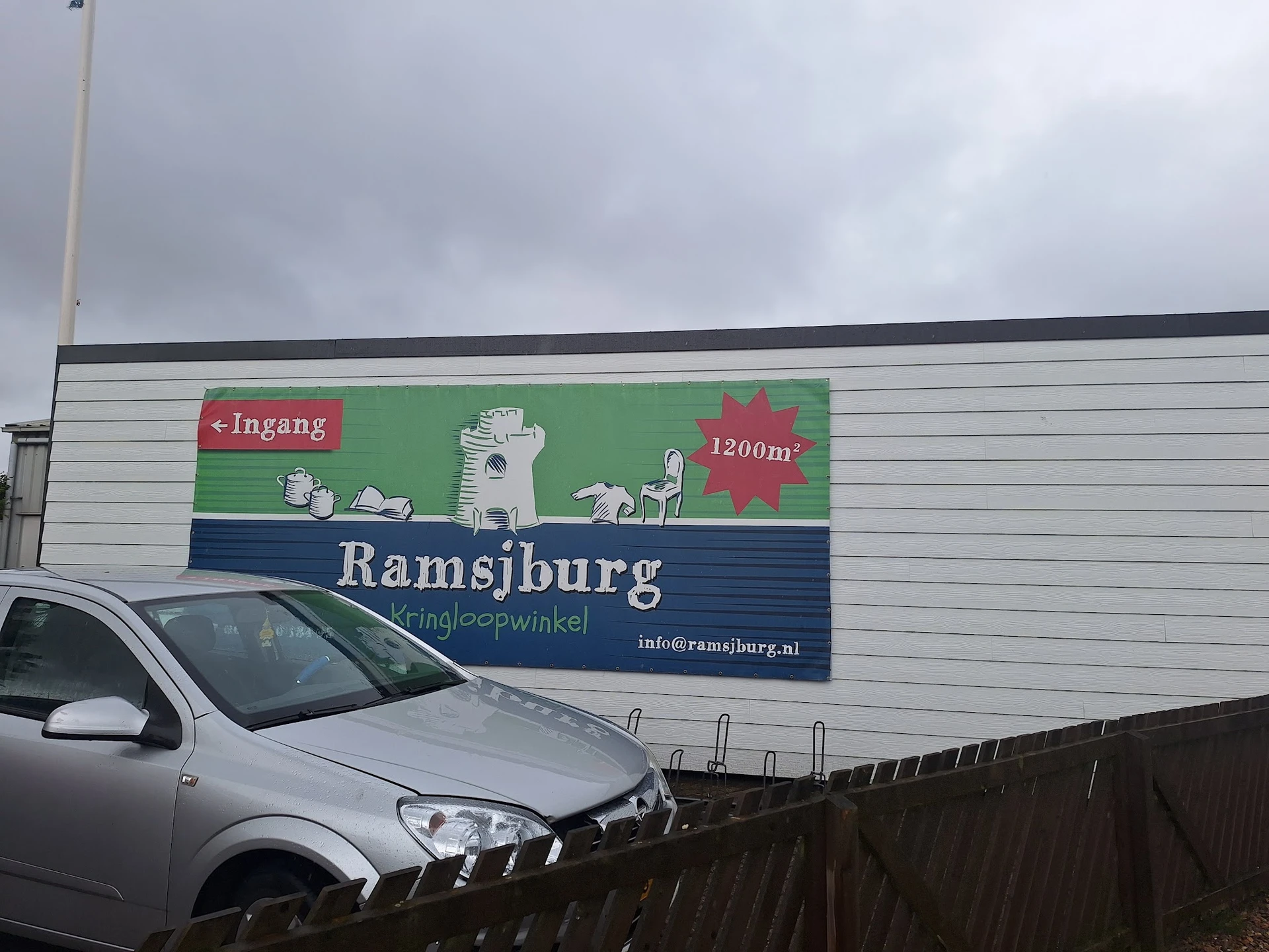 Gevel van kringloopwinkel Ramsjburg met groene en blauwe belettering, 1200m² aangegeven, met auto geparkeerd ervoor