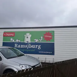 Gevel van kringloopwinkel Ramsjburg met groene en blauwe belettering, 1200m² aangegeven, met auto geparkeerd ervoor