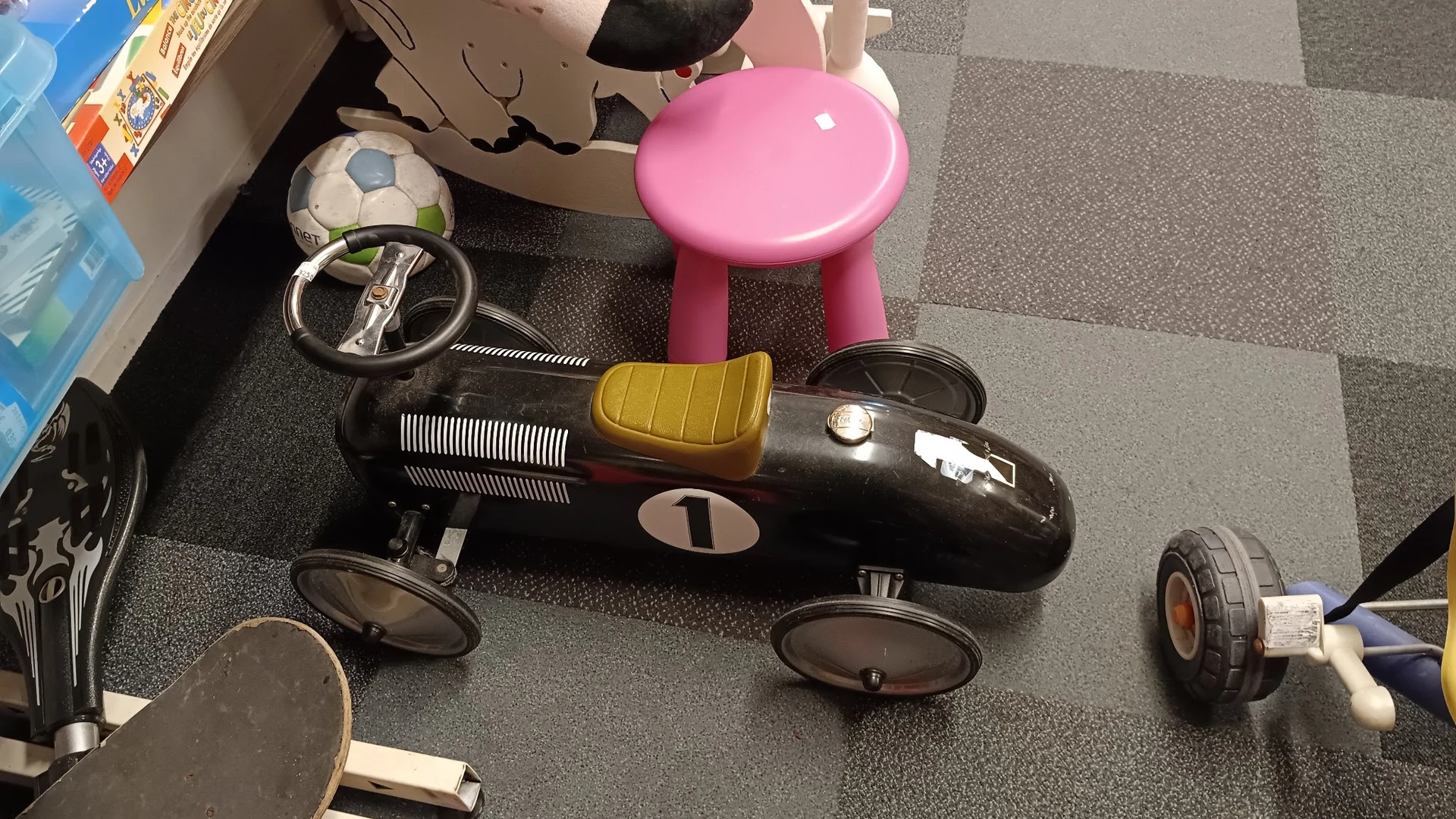 Verzameling speelgoedauto's en kinderspeelgoed op grijze vloer, waaronder zwarte racewagen met geel detail en roze kinderauto