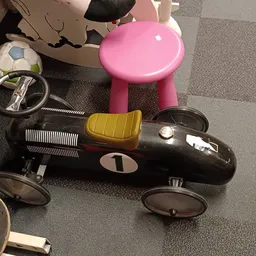 Verzameling speelgoedauto's en kinderspeelgoed op grijze vloer, waaronder zwarte racewagen met geel detail en roze kinderauto