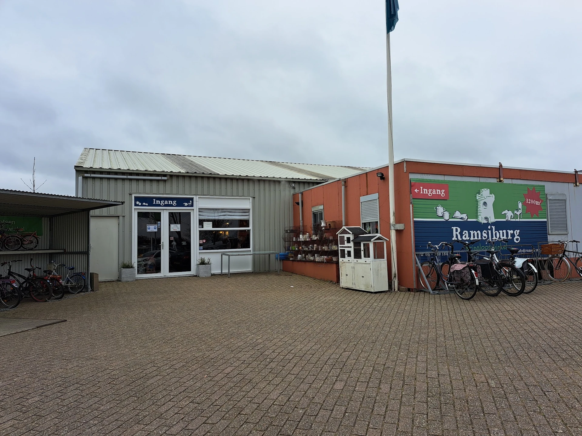 Exterieur van kringloopwinkel Ramsjburg met witte en rode bedrijfsgebouwen, parkeerplaats met fietsen, en bedrijfsnaam op gev