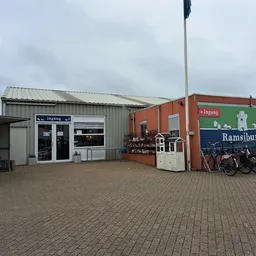 Exterieur van kringloopwinkel Ramsjburg met witte en rode bedrijfsgebouwen, parkeerplaats met fietsen, en bedrijfsnaam op gev