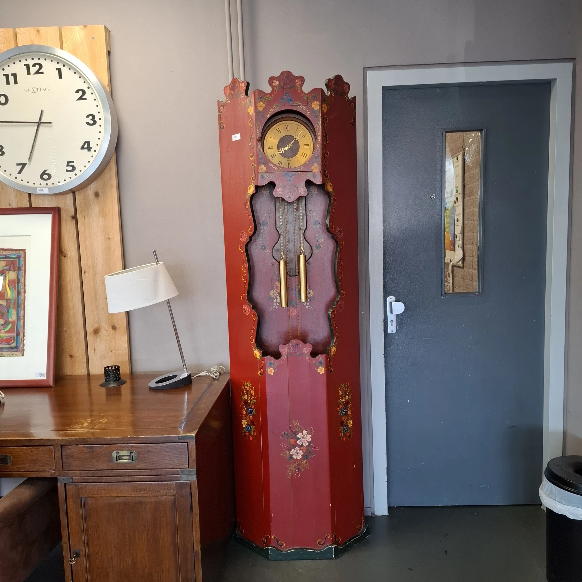 Winkelshoek met houten bureau, bureaulamp, wandklokken en vintage staande klok tegen rode muur