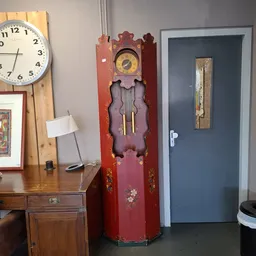 Winkelshoek met houten bureau, bureaulamp, wandklokken en vintage staande klok tegen rode muur