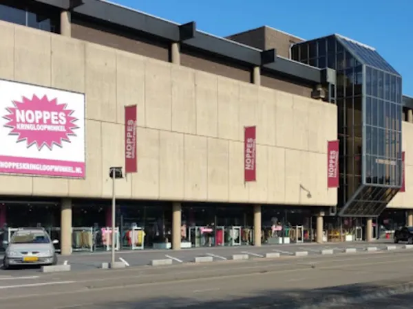 Gevel van kringloopwinkel met beige/taupe gekleurde voorgevel, roze NOPPES-logo en signage, grote ramen op begane grond, donk
