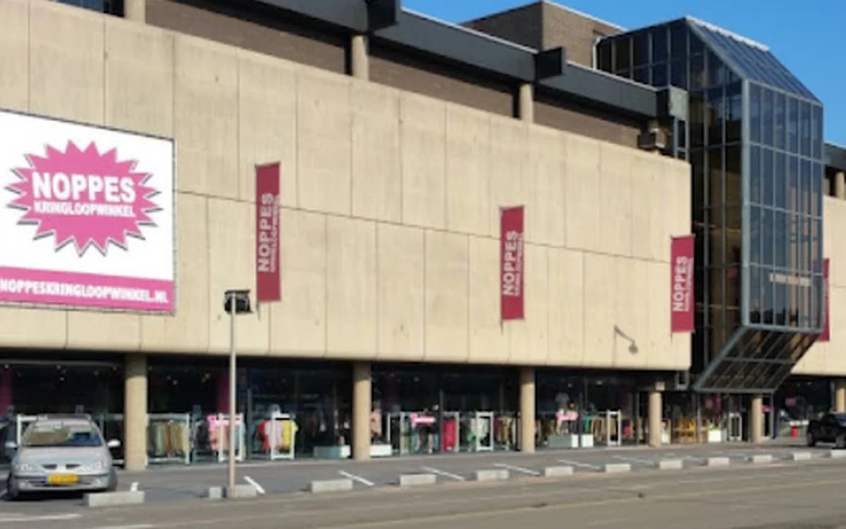 Gevel van kringloopwinkel met beige/taupe gekleurde voorgevel, roze NOPPES-logo en signage, grote ramen op begane grond, donk