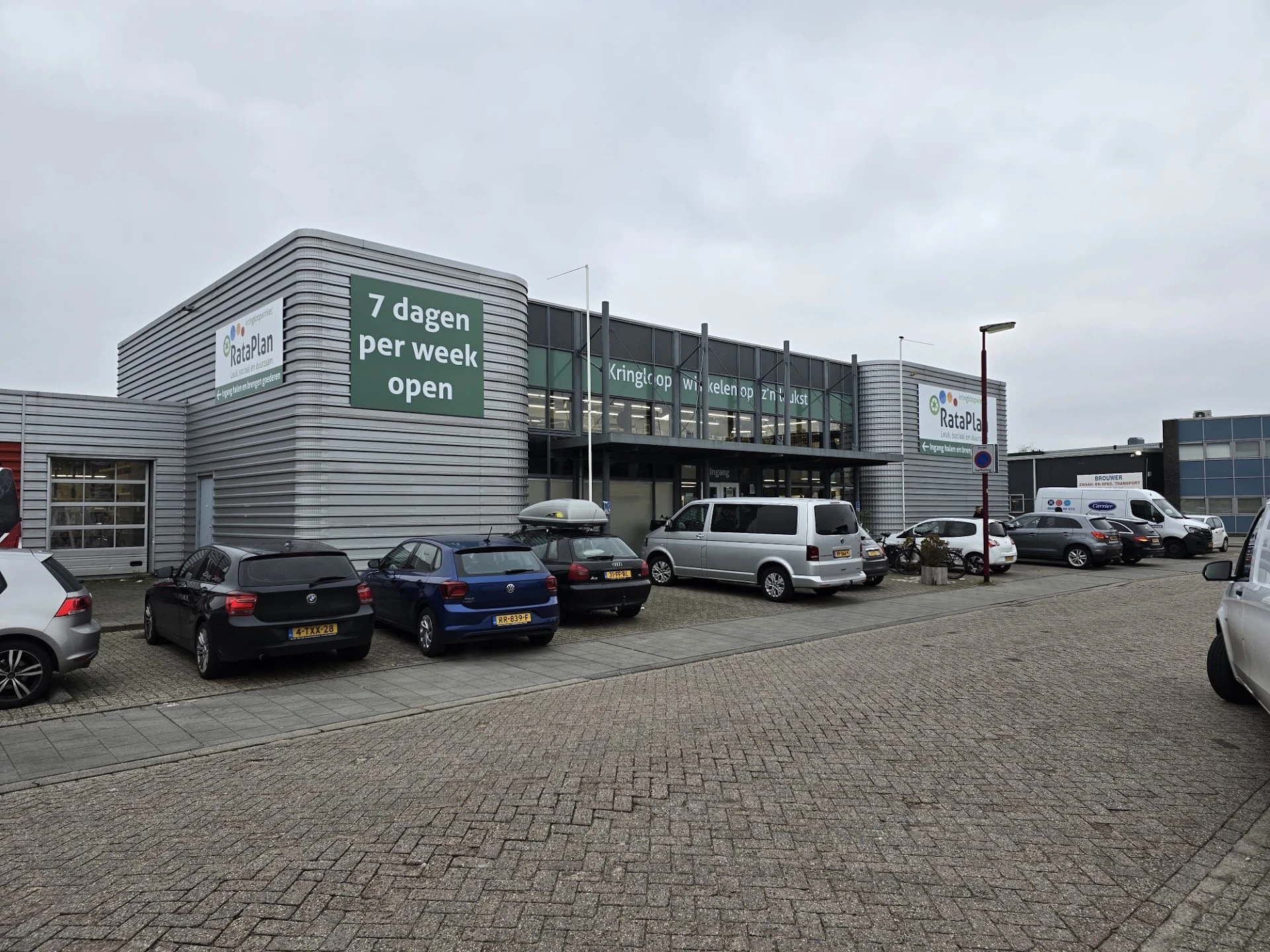 Grijze gevel van kringloopwinkel met witte letters 'Kringloop' en 'Kringloop 7 dagen per week open' op donkere ochtend, parke