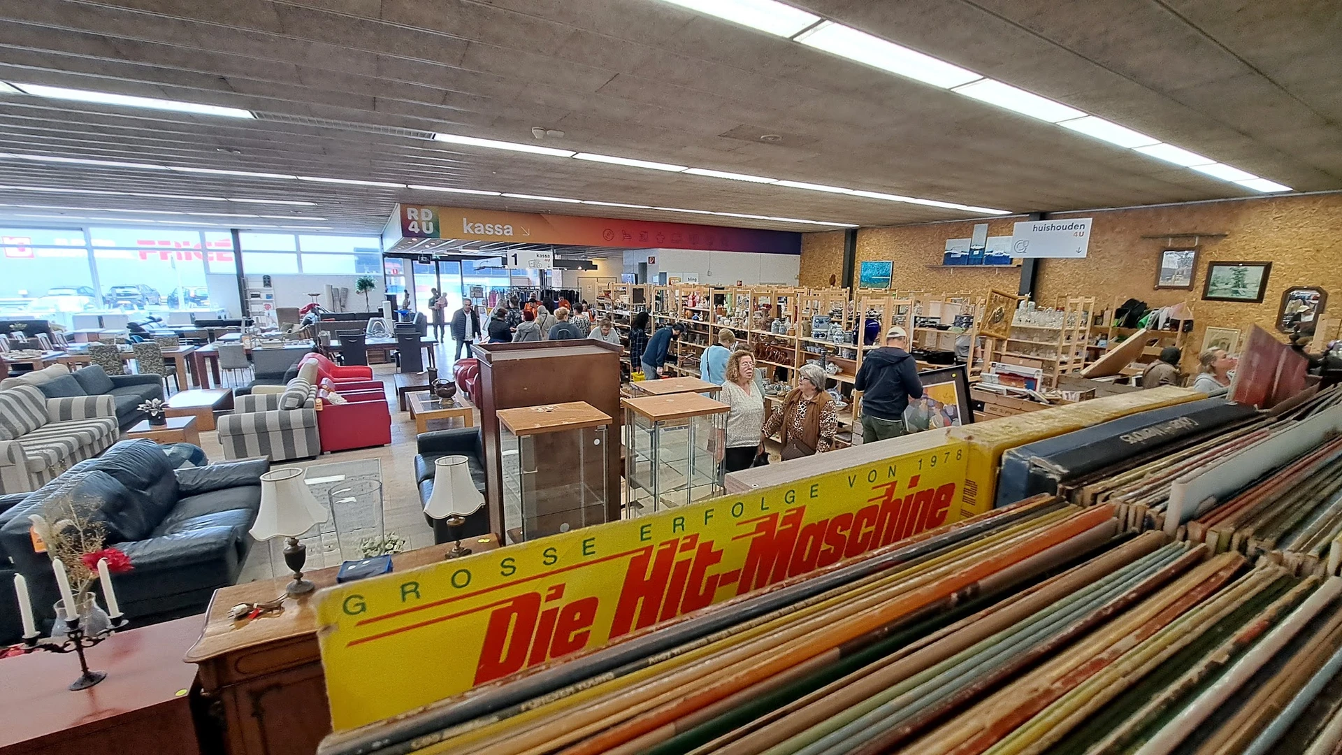 Winkelvloer van kringloopwinkel met diverse meubels, stellingen en bezoekers, kassengebied zichtbaar op achtergrond