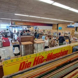 Winkelvloer van kringloopwinkel met diverse meubels, stellingen en bezoekers, kassengebied zichtbaar op achtergrond