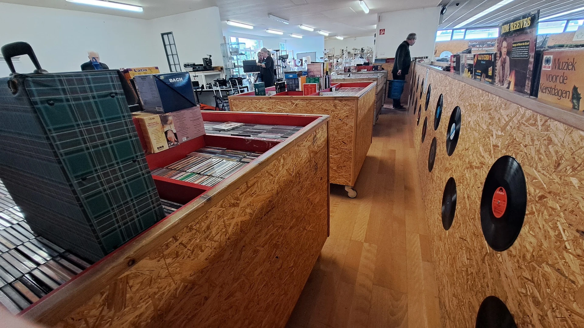 Winkelvloer met houten displaykasten en rekken vol tweedehands artikelen, personeel in het achtergrond