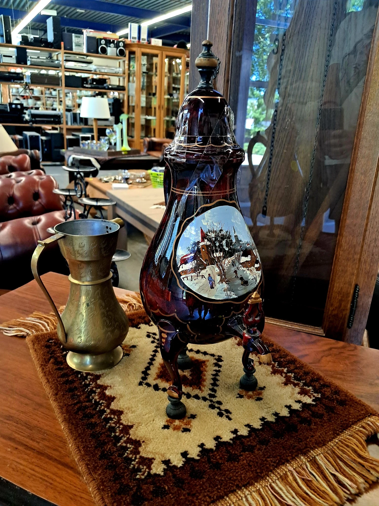 Close-up van een donkerrode glazen fles met decoratief winterlandschapbeeld en een gouden koperen kruik op houten tafel