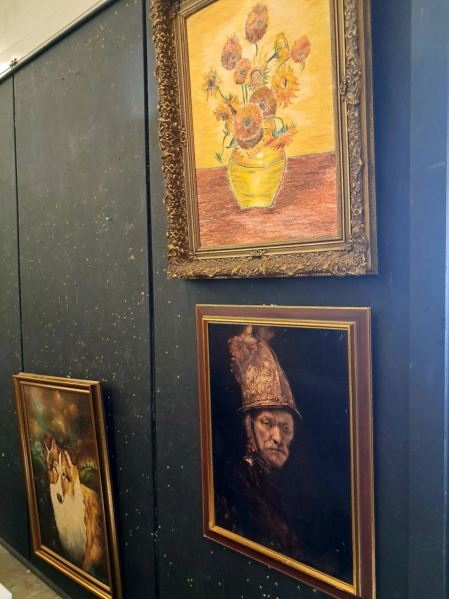 Ingelijste schilderijen aan een donkerblauwe wand, waaronder een stilleven met zonnebloemen en een portret met hoofdtooi