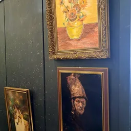 Ingelijste schilderijen aan een donkerblauwe wand, waaronder een stilleven met zonnebloemen en een portret met hoofdtooi