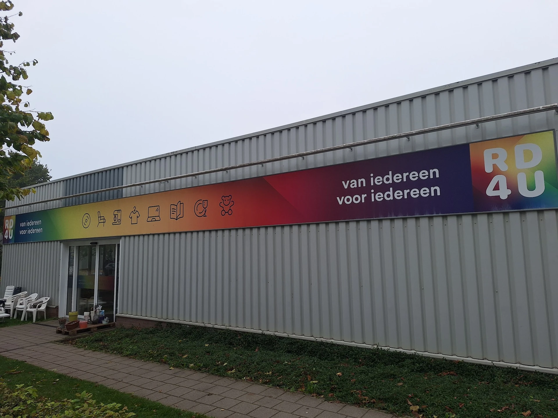 Grijze metalen voorgevel van kringloopwinkel met gele en rode bewegwijzering, ingang links, groen terrein ernaast