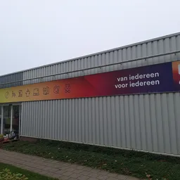 Grijze metalen voorgevel van kringloopwinkel met gele en rode bewegwijzering, ingang links, groen terrein ernaast
