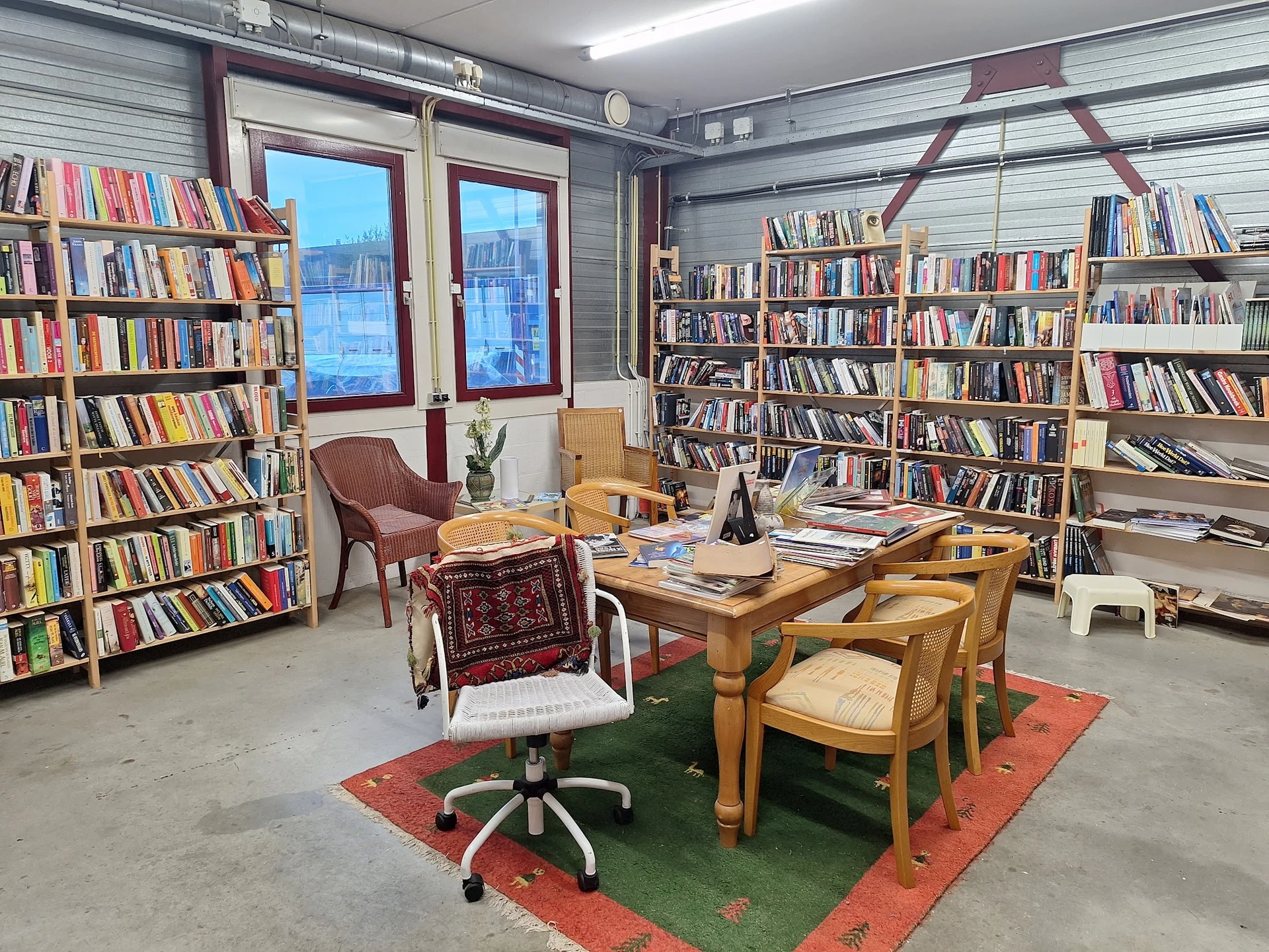 Leesruimte in kringloopwinkel met boekenkasten, houten tafel, stoelen en uitzicht op ramen