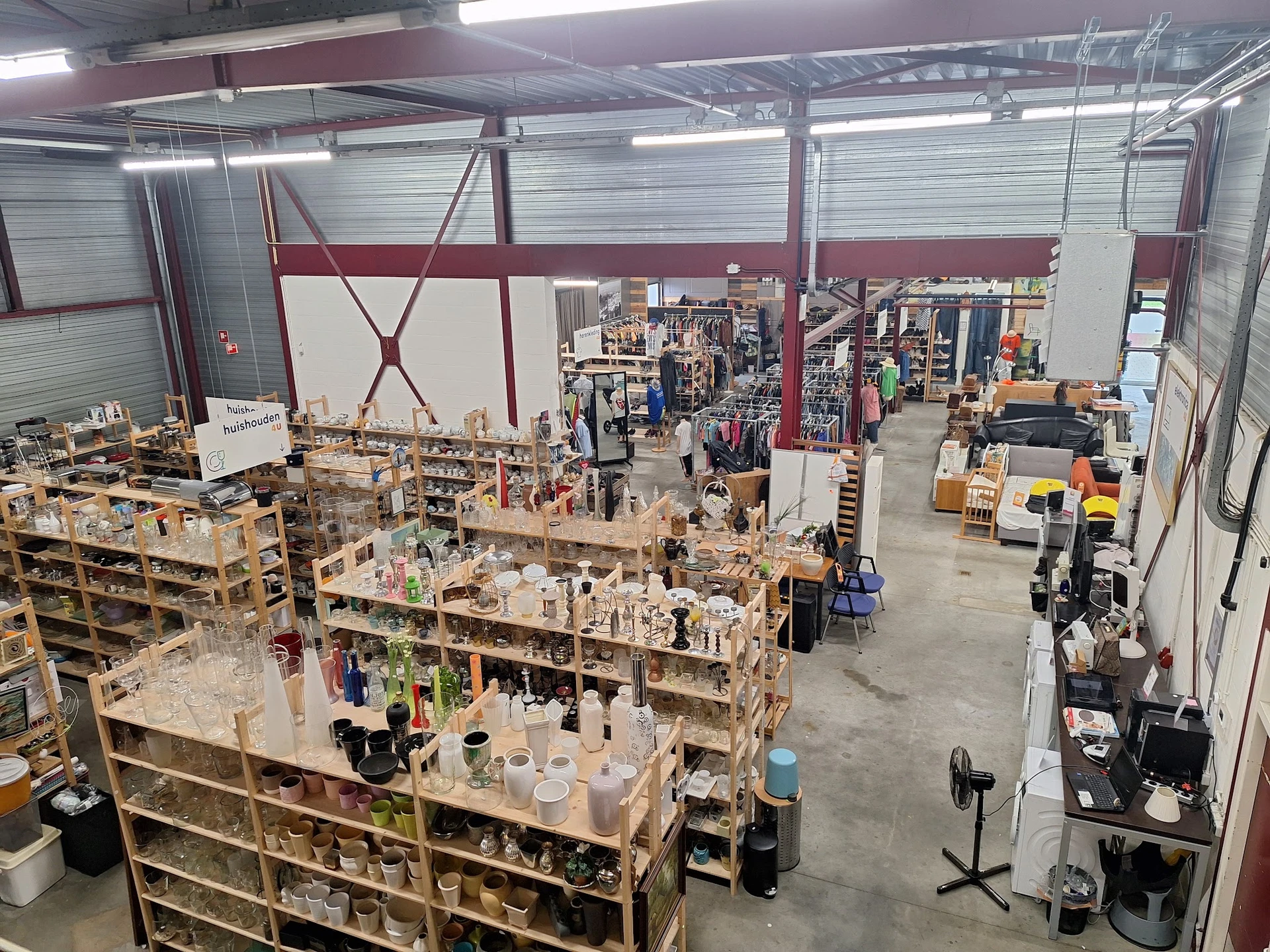 Overzicht van de kringloopwinkel met houten stellingen vol spullen, klanten kijken rond en verschillende afdelingen in de rui