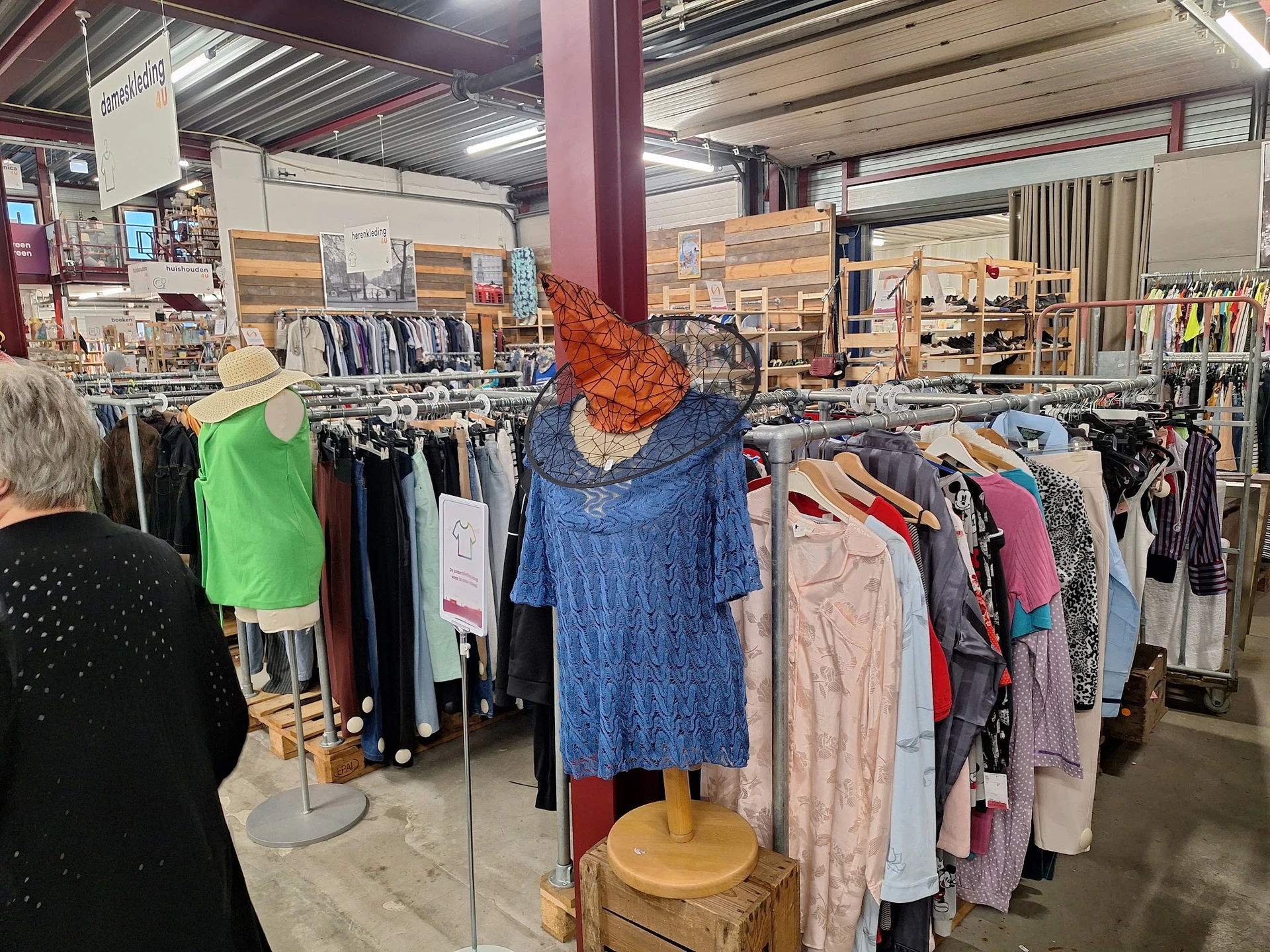 Rekken met tweedehands kleding, waaronder een blauwe jurk op een paspop met oranje hoed in het centrum van de winkel