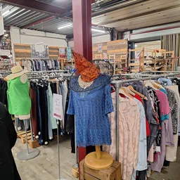 Rekken met tweedehands kleding, waaronder een blauwe jurk op een paspop met oranje hoed in het centrum van de winkel