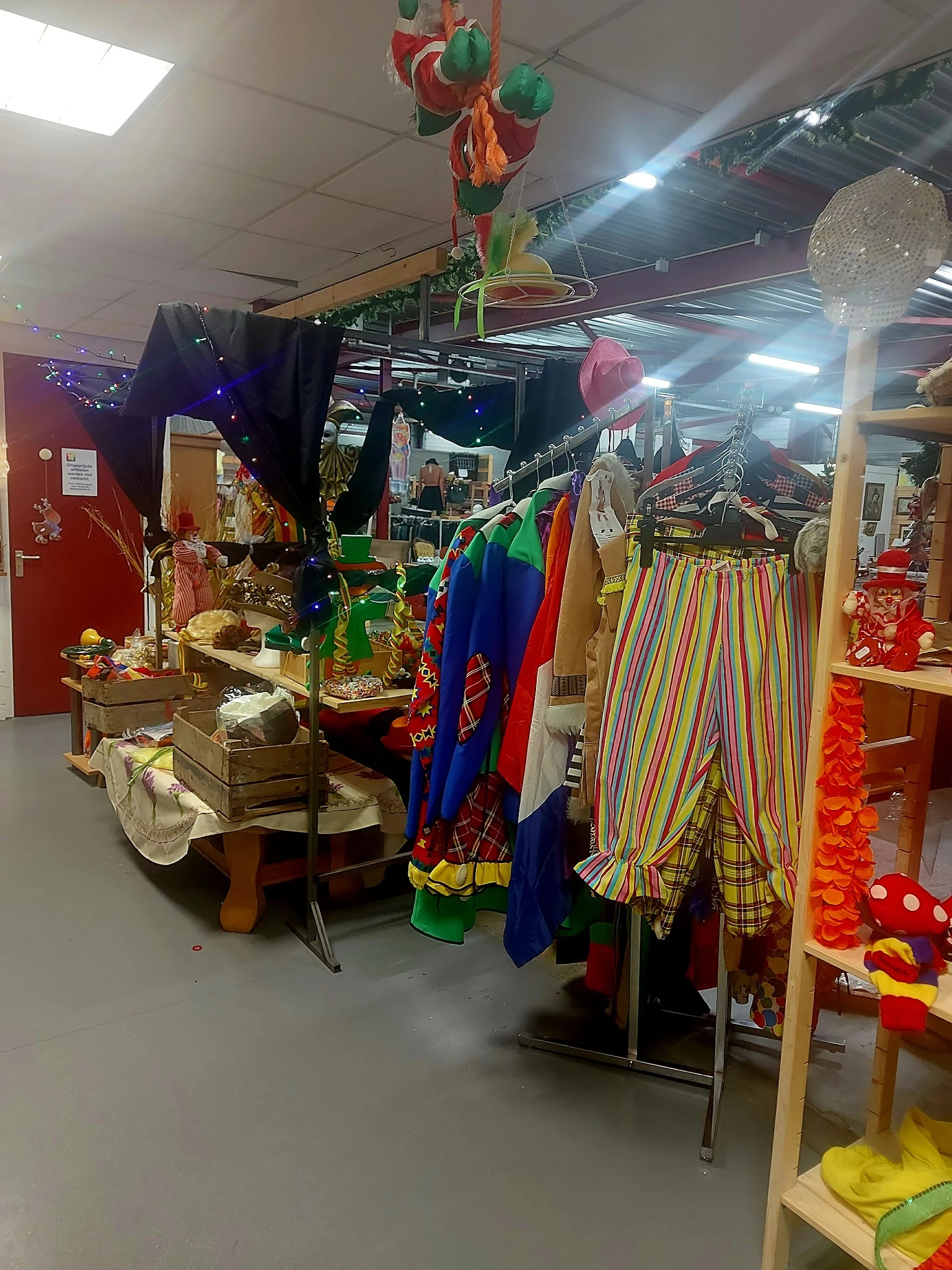 Rekken met kleurrijke carnavalskostuums en -attributen, waaronder clownspakken en regenbooggestreepte broeken in de kringloop