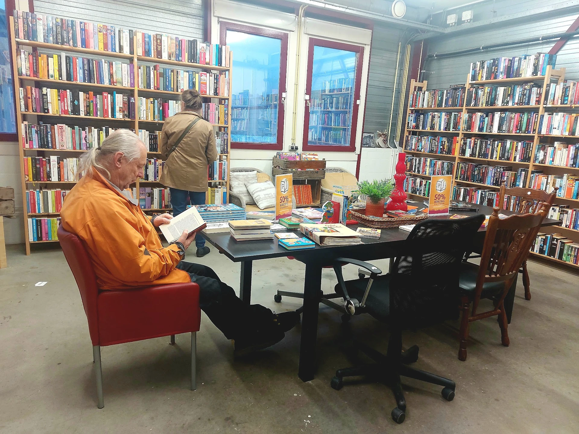 Boekenkasten langs de wanden van een kringloopwinkel met leeshoek waar een man in oranje jasje aan een tafel zit