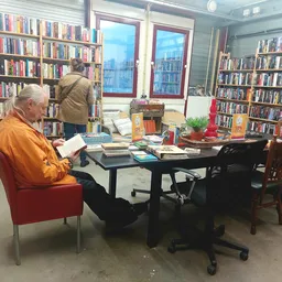 Boekenkasten langs de wanden van een kringloopwinkel met leeshoek waar een man in oranje jasje aan een tafel zit
