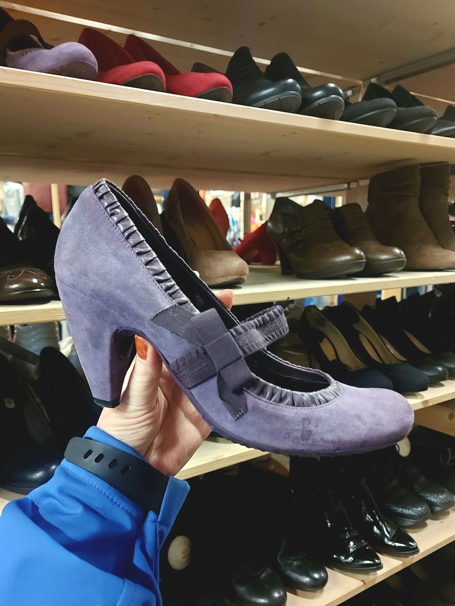 Close-up van een paarse suède pump met zwart accent, gehouden in hand tegen achtergrond van schoenenrekken