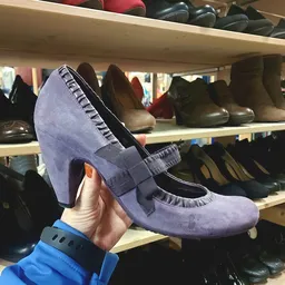 Close-up van een paarse suède pump met zwart accent, gehouden in hand tegen achtergrond van schoenenrekken