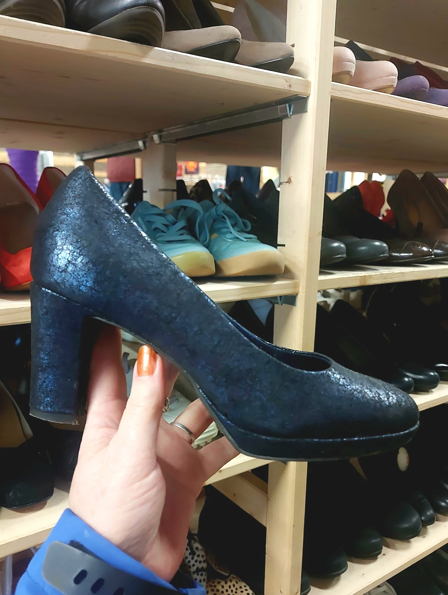 Close-up van een donkerblauwe glitterpump met blok hak, in hand tegen achtergrond met schoenen op winkelrekken