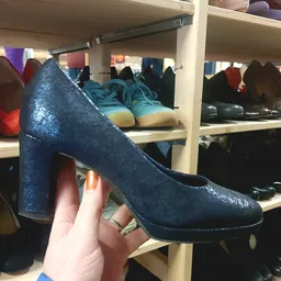 Close-up van een donkerblauwe glitterpump met blok hak, in hand tegen achtergrond met schoenen op winkelrekken