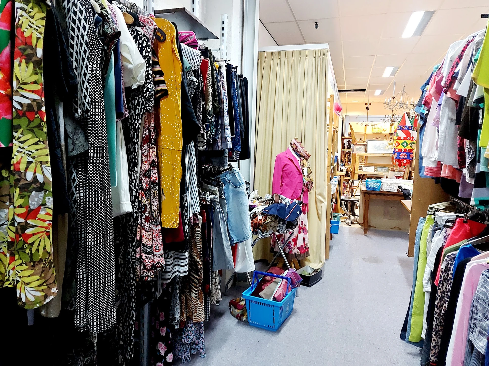 Kringloopwinkel met rekken vol tweedehands kleding, stellingen met spullen op de achtergrond en een winkelpad.