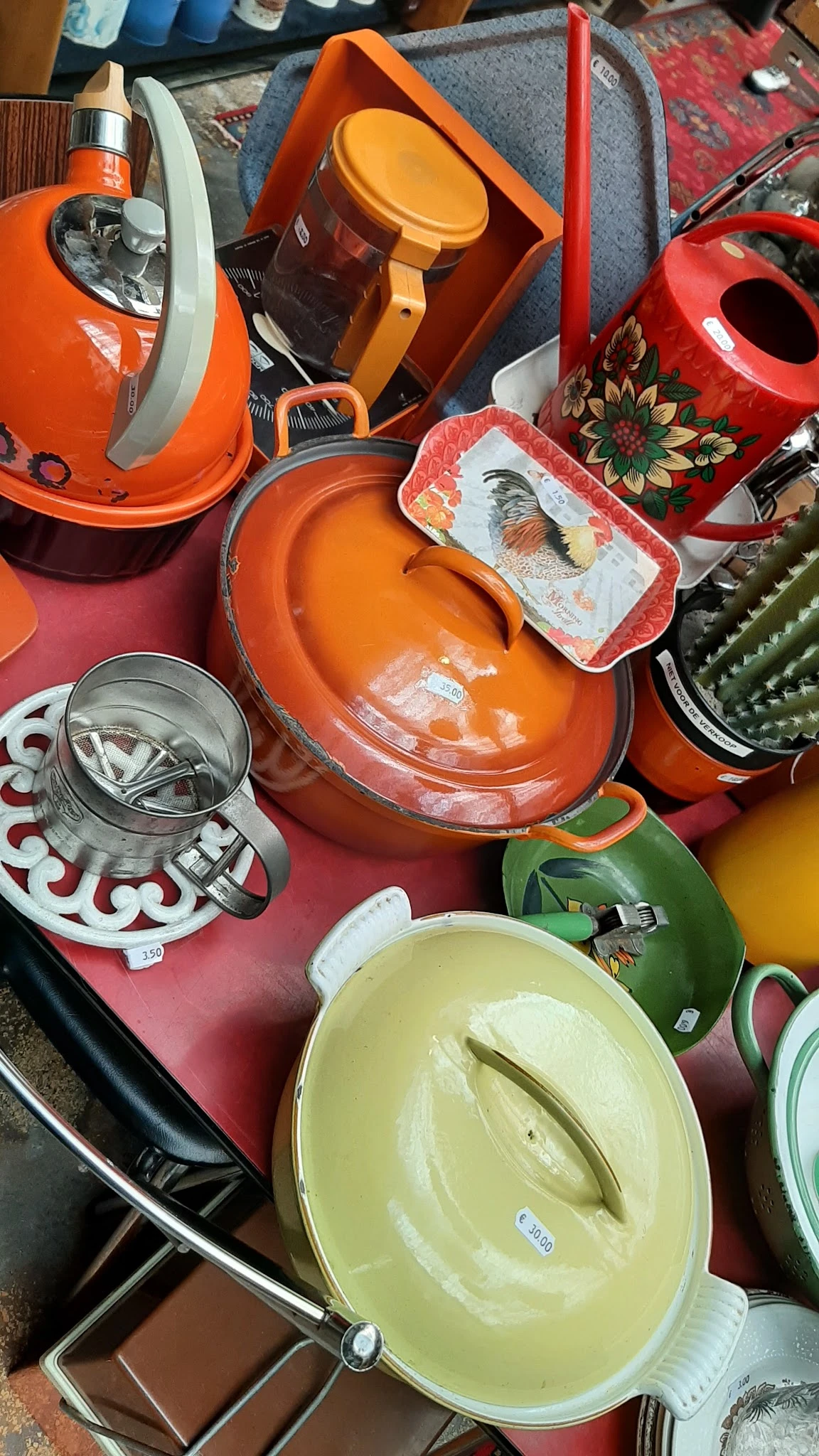 Verzameling kleurrijke huishoudelijke artikelen: oranje pannen met deksels, sieves, en decoratieve borden op roze ondergrond