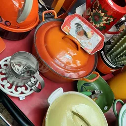 Verzameling kleurrijke huishoudelijke artikelen: oranje pannen met deksels, sieves, en decoratieve borden op roze ondergrond