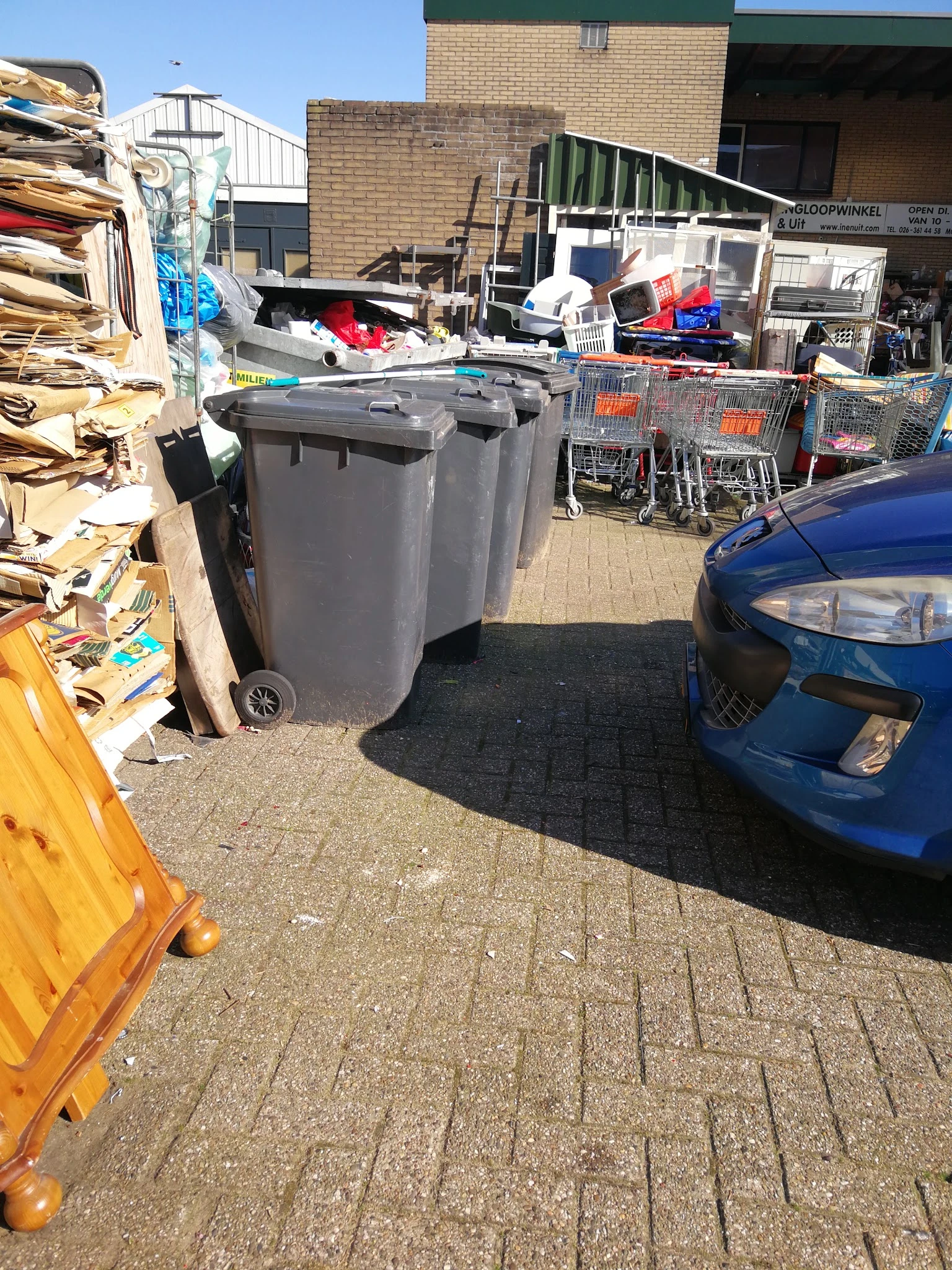 Parkeerterrein met grijze afvalcontainers, winkelwagen en blauwe auto naast ingang kringloopwinkel