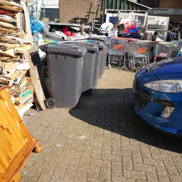 Parkeerterrein met grijze afvalcontainers, winkelwagen en blauwe auto naast ingang kringloopwinkel
