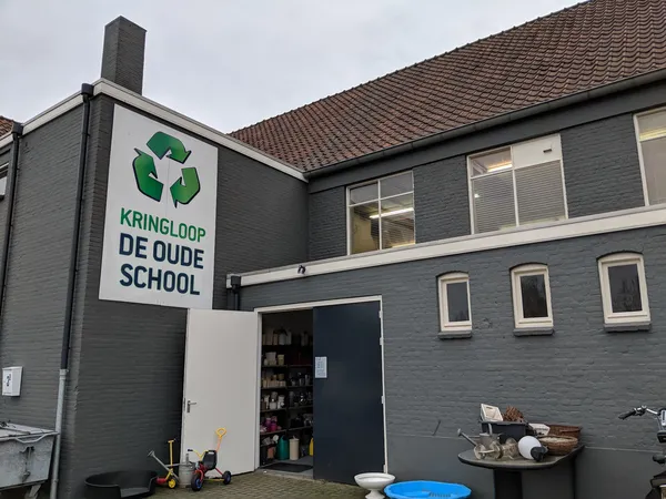 Grijze gevel van kringloopwinkel met wit bordje met groen recyclingsymbool en naam, ingang met donkerblauwe deur en ramen