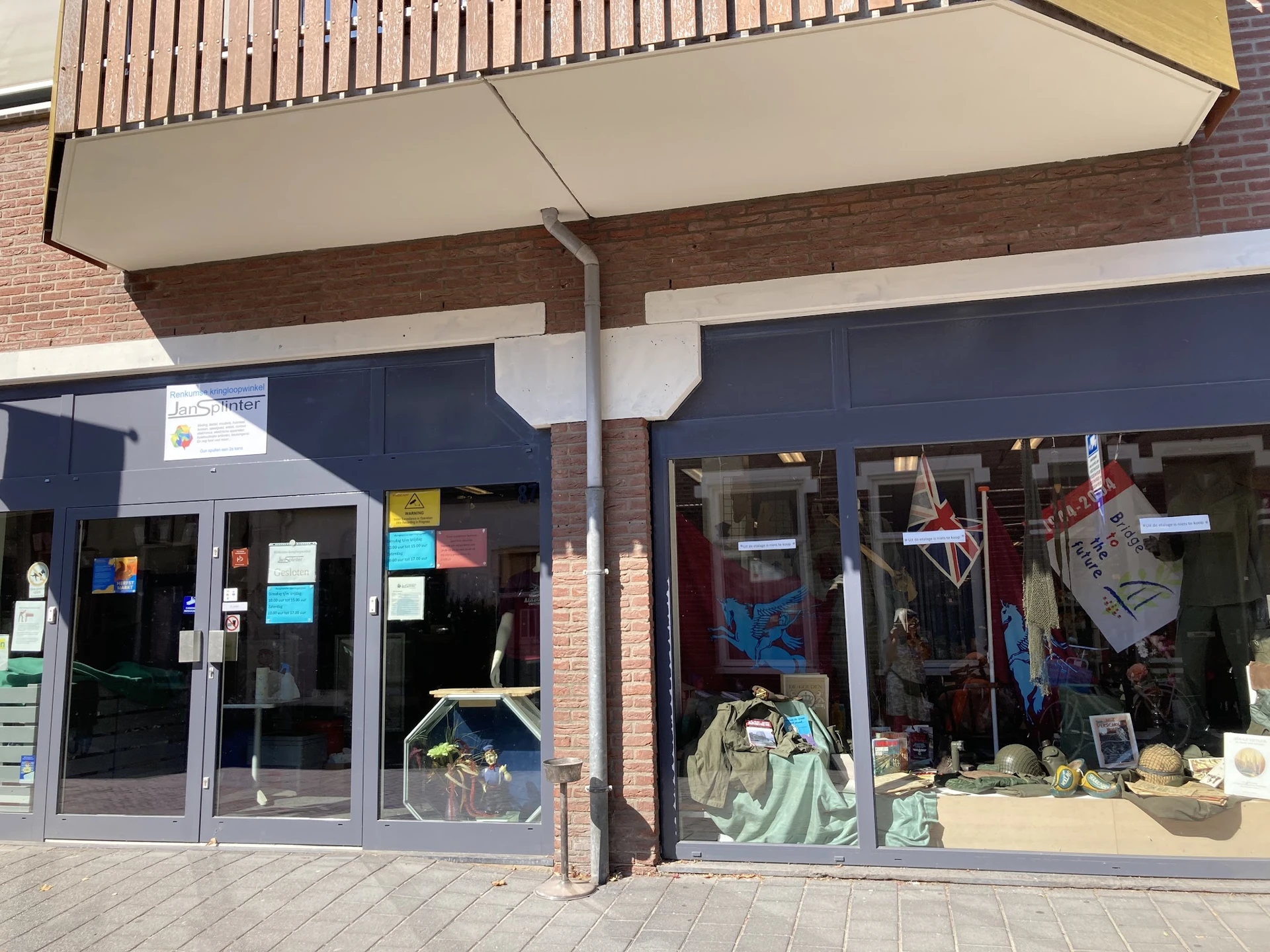 Voorkant van kringloopwinkel met grote ramen, donkerblauwe deuren en uitgestalde spullen zichtbaar achter het glas