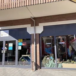 Voorkant van kringloopwinkel met grote ramen, donkerblauwe deuren en uitgestalde spullen zichtbaar achter het glas