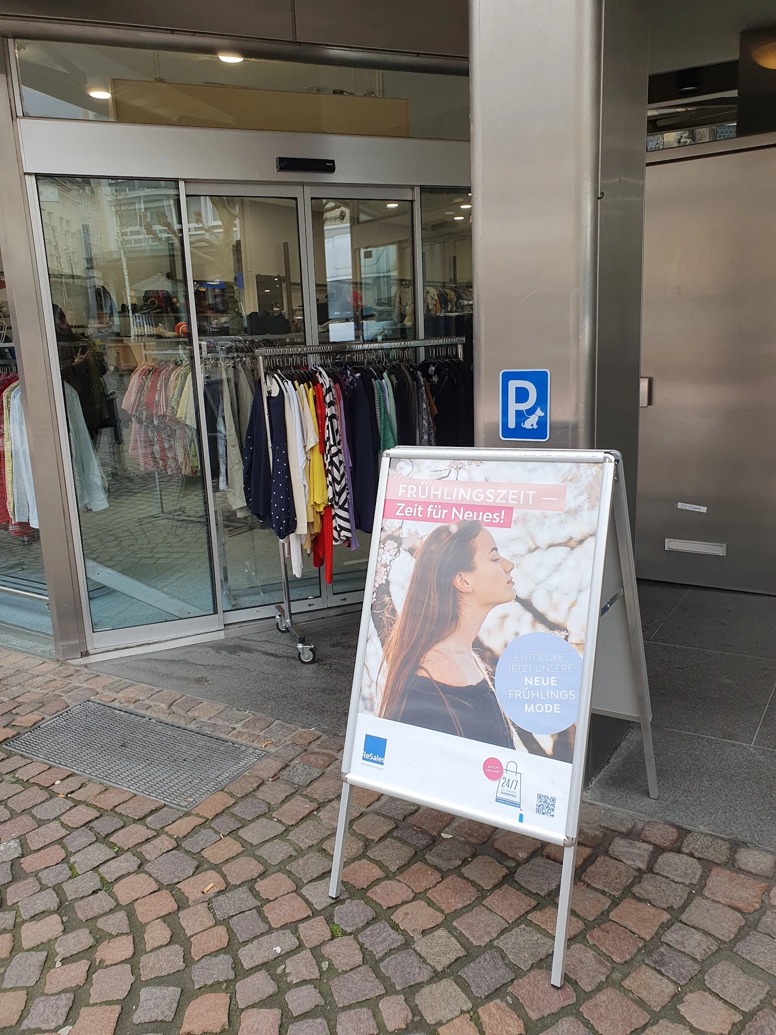 Winkelpui met glazen deuren en tweedehands kleding aan rekken, parkeerplaatsbord en promotiebord op straat